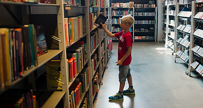 Ett barn står vid en lång bokhylla med böcker på ett bibliotek. Hen drar ut en bok.