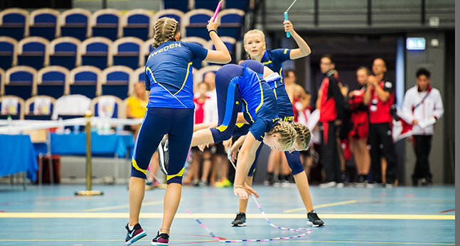 Fyra tjejer i blått och gult som är på golvet i en sporthall. Två snurrar de båda repen, två hoppar mitt i repen. De står på händerna, fast händerna är lyfta från marken mitt i ett hopp.