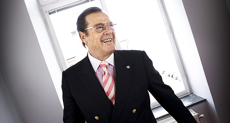 Roger Moore står framför ett fönster och ler
