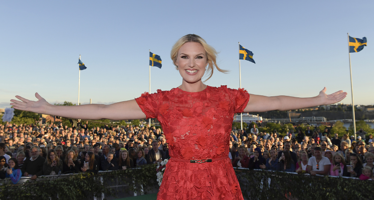 Sanna Nielsen i röd klänning står på scenen på Skansen. Hon sträcker ut armarna åt sidorna och ser glad ut. Bakom henne ser man publiken, en blå himmel och två svenska flaggor som vajar.