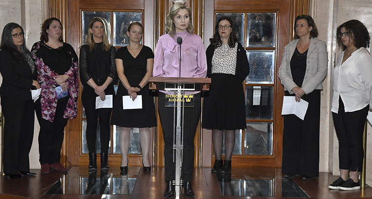 Ebba Bush Thor står vid en pulpet och pratar i en mikrofon. Bakom henne står flera politiker uppradade. De är alla kvinnor och ser allvarliga ut.