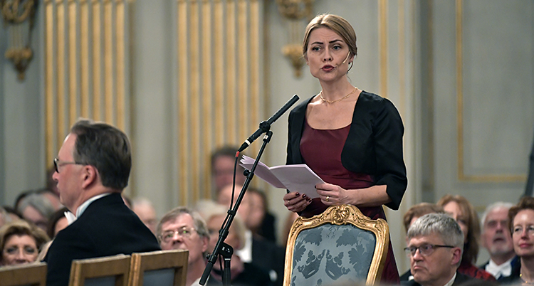 Jayne Svenungsson håller tal. Hon står bakom sin stol och pratar in i en mikrofon. I händerna håller hon ett papper. Stolen har en ram av guld. Den är också klädd i blått tyg.