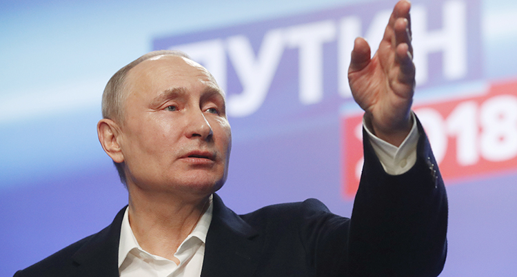 Vladimir Putin står på en scen och pratar. Han pekar med sin hand. Han har en vit skjorta och en mörk kavaj.