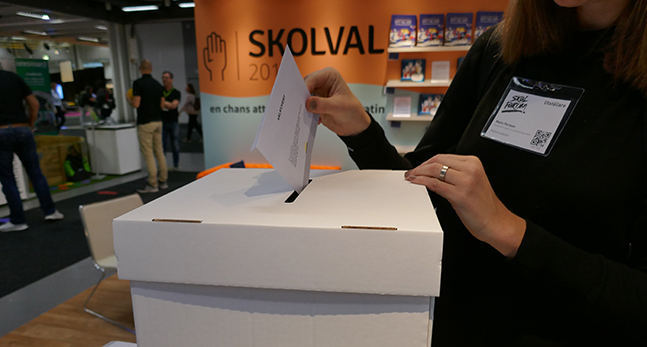 En låda står i ett bibliotek. En person stoppar ett valkuvert i lådan.