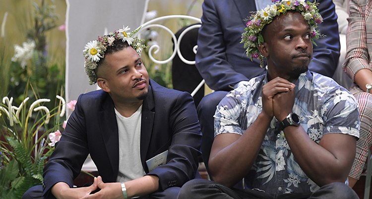 De sitter bredvid varandra . Erik i kavaj och vit t-shirt, Kodjo i mönstrad skjorta. Båda har blomsterkrans.