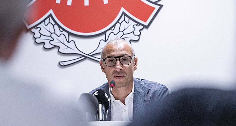 Henrik Larsson sitter vid en skrivbord bakom ett par mikrofoner. Han ser ledsen ut.