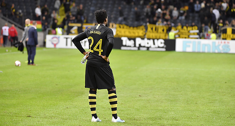 En spelare i AIK står på planen och deppar efter att matchen är slut. Han har svarta kläder med gula detaljer.