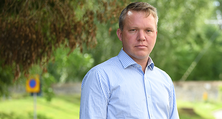 En dyster man står i rutig vit skjorta. Han tittar in i kameran och är utomhus.