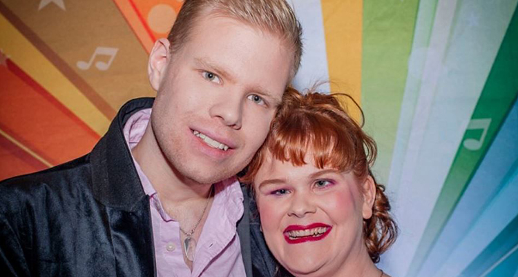 En blond man och en rödhårig kvinna ler och tittar in i kameran. De står mot en vägg i regnbågens alla färger.