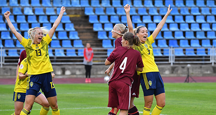 Två svenska spelare lyfter armarna i luften och jublar efter ett mål. De har gula tröjor och blå shorts. Några lettiska spelare syns också i bilden. De har röda dräkter. Läktaren i bakgrunden har blå säten.