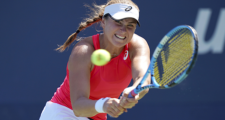 Rebecca Peterson spelar tennis och slår till en boll i en match. Hon har på sig ett orange linne och en vit keps.