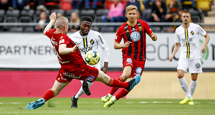 En fotbollsmatch. Fyra spelare syns i bilden. En spelare i rött och en spelare i vitt kämpar om bollen. De andra två spelarna syns i bakgrunden.