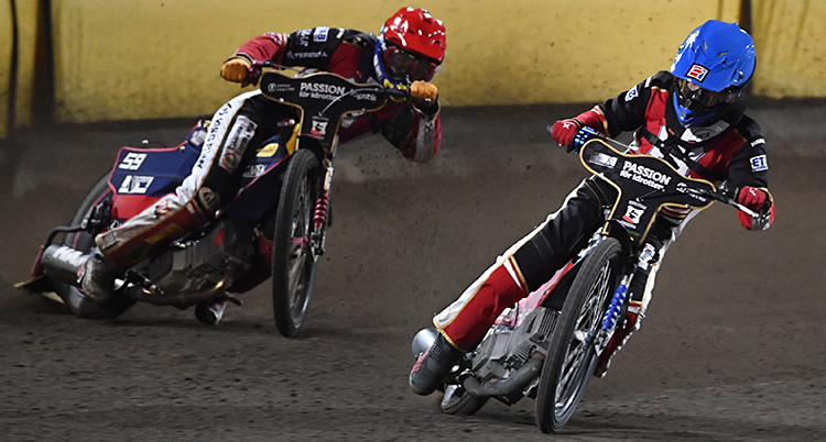 Två förare som kör speedway på en bana. De sladdar i en kurva. Föraren som leder har en blå hjälm och den andra har en röd hjälm.