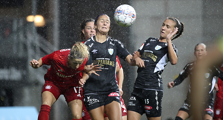 Tre spelare kämpar om bollen i en fotbollsmatch. Bollen är uppe i luften. Två spelare har svarta tröjor och en spelare har röd tröja.