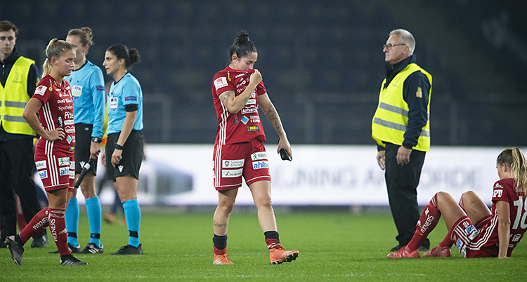 Tre spelare i laget Piteå ser ledsna ut. De är på en fotbollsplan. De har röda kläder på sig. Bredvid syns några domare och några funktionärer med reflexvästar.