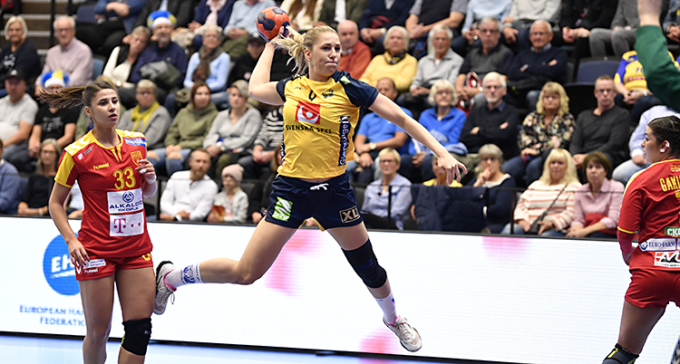 Vi ser en bild från en handbollsmatch. En kvinna hoppar upp. Hon har bollen i handen och laddar för ett skott. Hon har blå shorts och gul tröja. Bredvid henne syns två motståndare. De har röda kläder. I bakgrunden syns publiken.