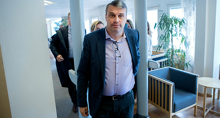 En man har precis gått igenom en larmbåge i en domstol. Han har en lila skjorta och en mörk kavaj. Han har håret kammat i en sidbena. Han har mörkt hår med ett grått parti i håret längst fram.