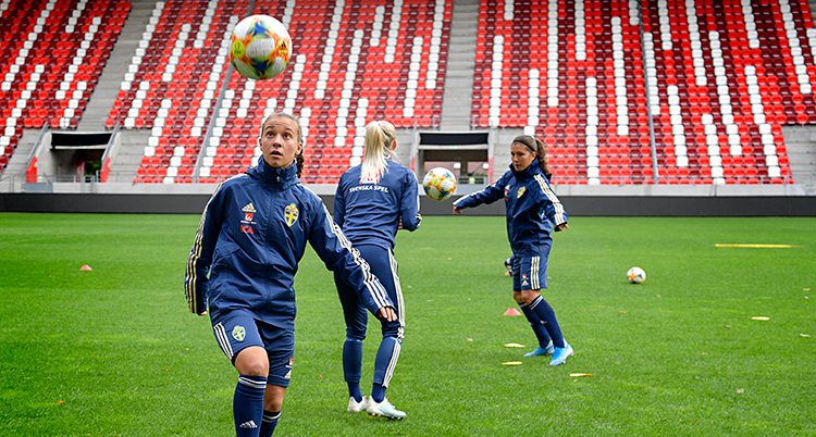 En bild från en träning med svenska landslaget i fotboll, damer. Tre spelare är på en fotbollsarena med tomma läktare. Spelarna har träningsoveraller på sig och tränar med bollar.