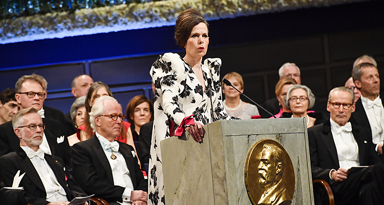 Sara Danius står i en talarstol i en vit klänning med svart mönster. Bakom henne sitter herrar i frack.