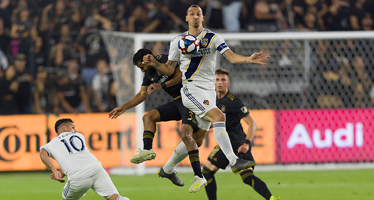 Bilden visar en match i fotboll. Zlatan Ibrahimovic hoppar upp i luften och tar bollen på bröstet. Runt om honom finns flera andra spelare.