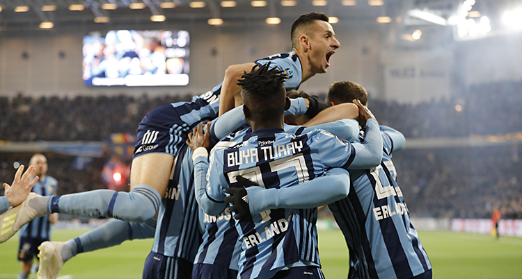 Bilden är tagen på Tele2 Arena i Stockholm. Flera spelare i Djurgården kramar varandra. De har tröjor som är randiga i mörkblått och ljusblått. I bakgrunden syns den gröna mattan och längre bort läktaren med publiken