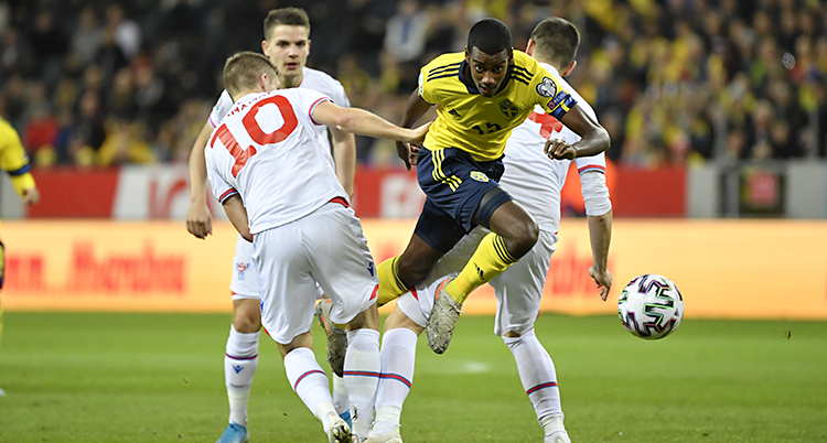 Från en fotbollsmatch mellan Sverige och Färöarna. Sveriges spelare Alexander Isak kämpar om bollen med en spelare från Färöarna. Isak har guld tröja och blå shorts. Den andra spelaren har vita kläder med röda siffror på ryggen.