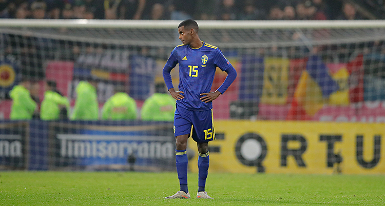 En fotbollsplan. Mitt i bilden står spelaren Alexander Isak. Han har Sveriges blå dräkt på sig. I bakgrunden syns ett fotbollsmål och publik.