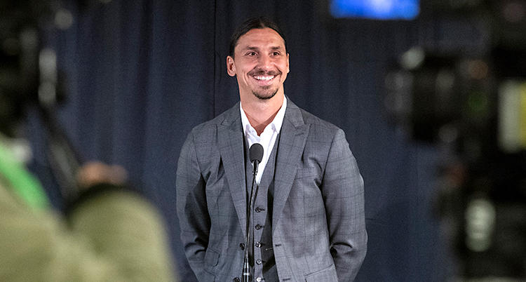 Fotbollsspelaren Zlatan Ibrahimovic står på en scen och pratar i en mikrofon. Han ler mot publiken. Han har på sig en grå kostym och vit skjorta.