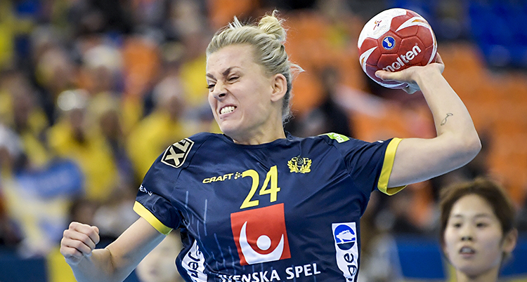 Bilden är från en match i handboll. Spelaren Nathalie Hagman laddar för ett skott. Hon har bollen i handen. Hon har blont hår som är uppsatt. Hennes tröja är blå med gula siffror.