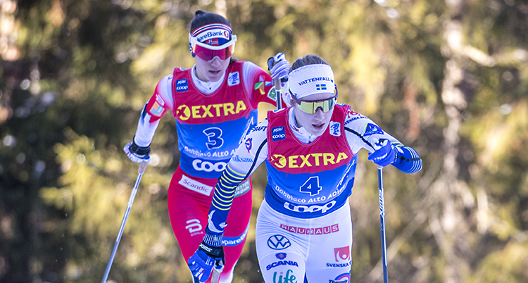 Tvåskidåkare åker nära varandra. Ebba är först och Heidi någon meter efter i spåret. Träd i bakgrunden.