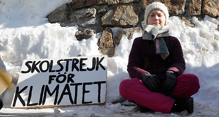 Svenska Greta Thunberg sitter på marken. Hon har vinterkläder på sig. Bakom henne syns en snödriva. Bredvid sig har hon ett plakat där det står Skolstrejk för klimatet.