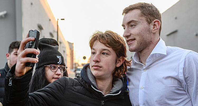 Fotbollsspelaren Dejan Kulusevski har vit skjorta. Han står på en gata i staden Turin. En person står bredvid honom. Personen tar en bild på dem tillsammans med sin mobiltelefon. Bredvid står en ung kvinna med en mössa som det står Juventus på.