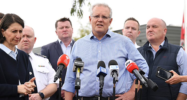 Australiens ledare Scott Morrison har en träff med journalister. Han står och pratar in i flera mikrofoner. Han har grått hår, glasögon och ljusblå skjorta. Runt honom står flera människor.