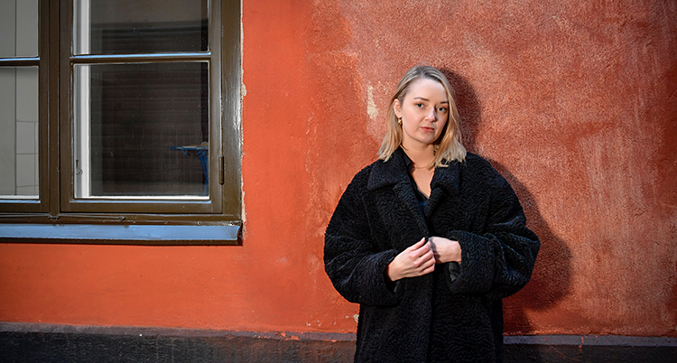 Elin Persson står mot en vägg. Den är röd. Elin har blont långt hår och en svart kappa på sig.