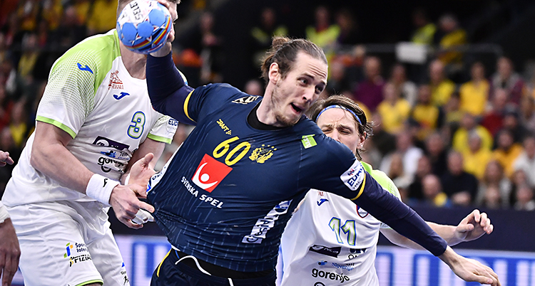 En svensk spelare hoppar för att skjuta i en match i handboll. Han har bollen i handen och mörkblå kläder på sig. Det andra laget har vita kläder.