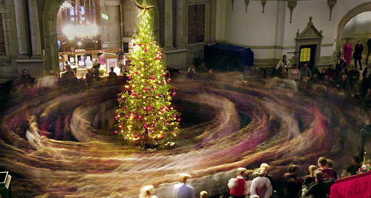 En stor julgran lyser i mitten av ett rum. Folk dansar runt granen. Bilden är tagen så att de som dansar är alldeles suddiga.