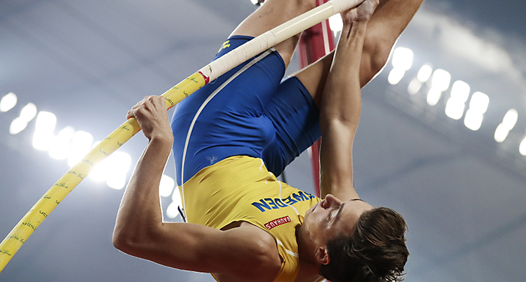 Bilden visar Armand Duplantis. Han hoppar stav. Han håller i sin stav och hänger upp och ner i luften. Han har gult linne och blå shorts.