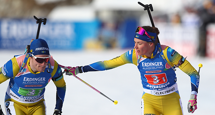 Det är en växling i stafett i skidskytte. Sebastian Samuelsson är till vänster. Hanna Öberg är till högre. Hon lägger sin hand på Sebastians axel. Båda har på sig Sveriges dräkter i gult och blått.