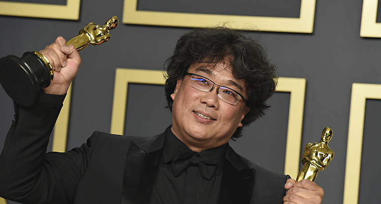 Bong Joon Ho håller upp en statyett med Oscars i varje hand. Han har glasögon och svart hår.