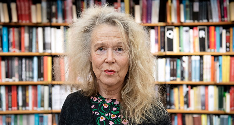 En kvinna med ljust hår. Håret är långt och stort. Hon är i ett bibliotek. Bakom syns hyllor med böcker.