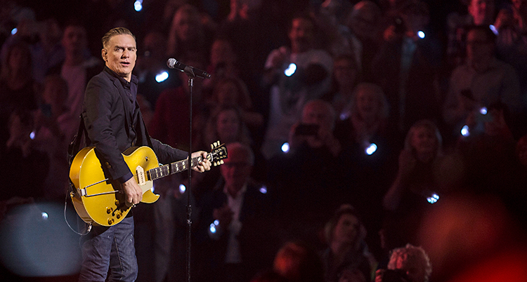 Bryan Adams står på en scen med sin gitarr. I bakgrunden syns publiken. Många håller upp sina mobiltelefoner som lyser.