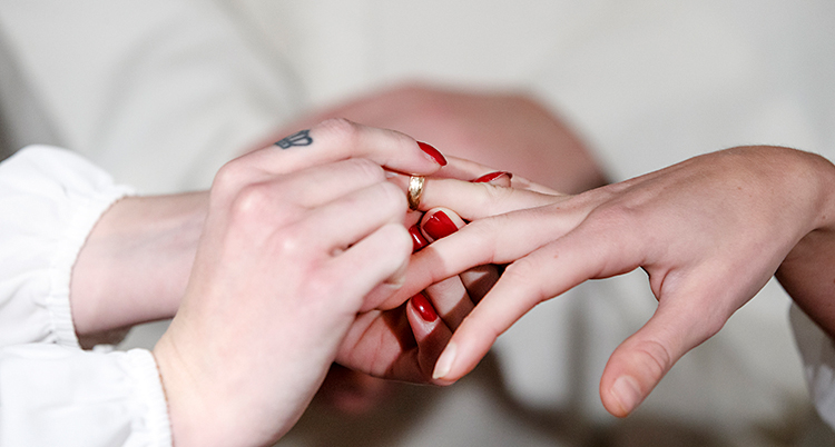 Bilden visar händerna från två personer. Den ena personen sätter en ring på fingret på den andra.