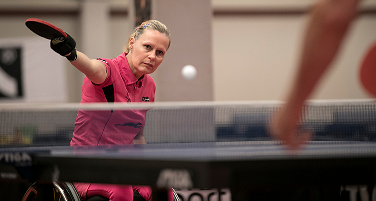 En kvinna som spelar bordtennis. Hon slår en boll över nätet. Hon sitter i en rullstol.