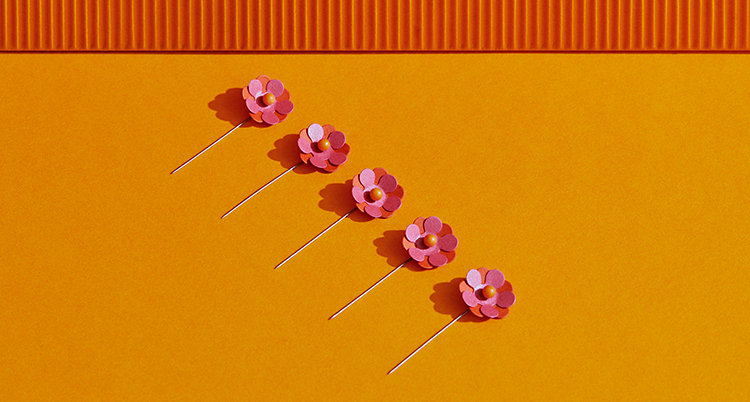 Fem stycken majblommor ligger på en orange bakgrund. Blommorna är rosa och orange. Alla blommor sitter på en liten pinne av metall.
