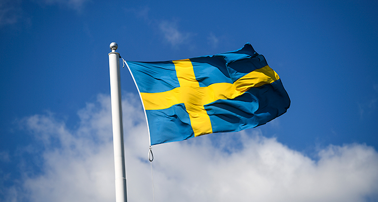 En svensk flagga vajar i vinden på en flaggstång. Himlen är blå med några moln.