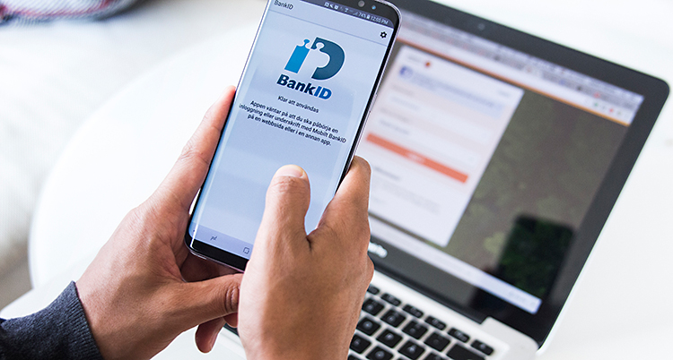 En person sitt med sin mobiltelefon. Personen loggar in på bank-id. I bakgrunden står en dator på ett bord.