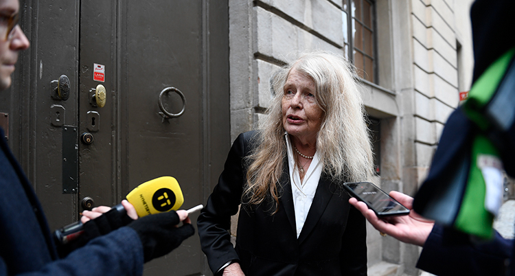 Hon har långt vitt hår och en svart kavaj. Journalister sticker fram mikrofoner