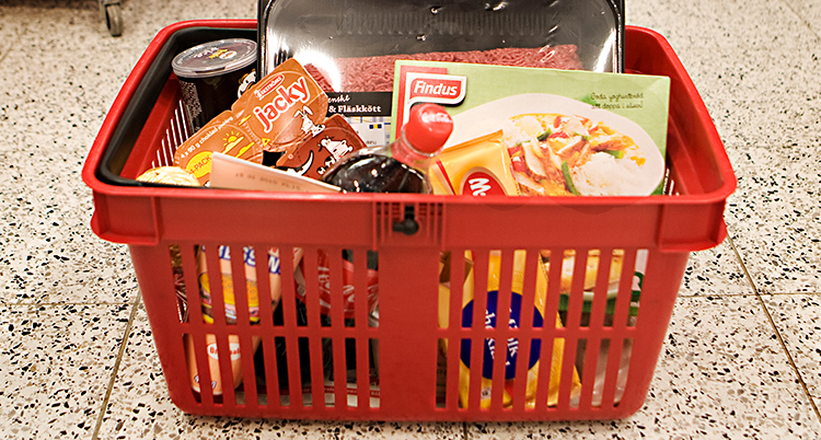 Korgen är röd. Den står på golvet. I korgen ligger massa varor. Bland annat en flaska med cola och en chokladkaka.