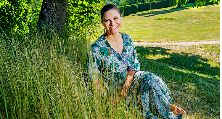 Kronprinsessan Victoria sitter i gräset. Hon ler och har en grönblå sommardräkt på sig.