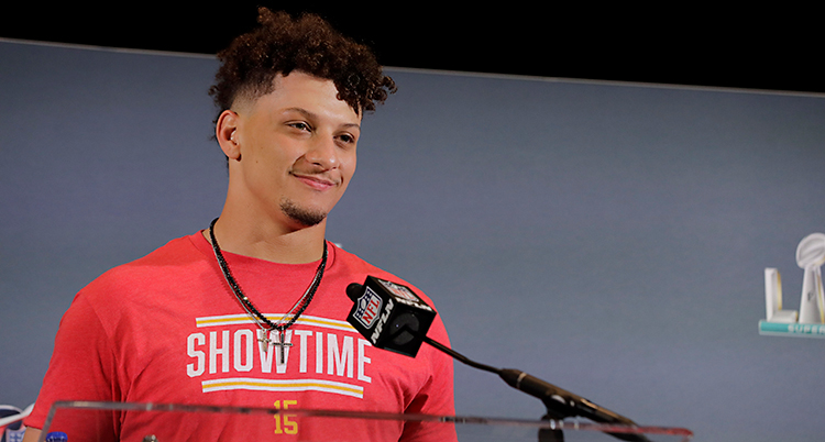 Patrick Mahomes står på en scen. Han pratar i en mikrofon. Han har en röd t-shirt på sig.